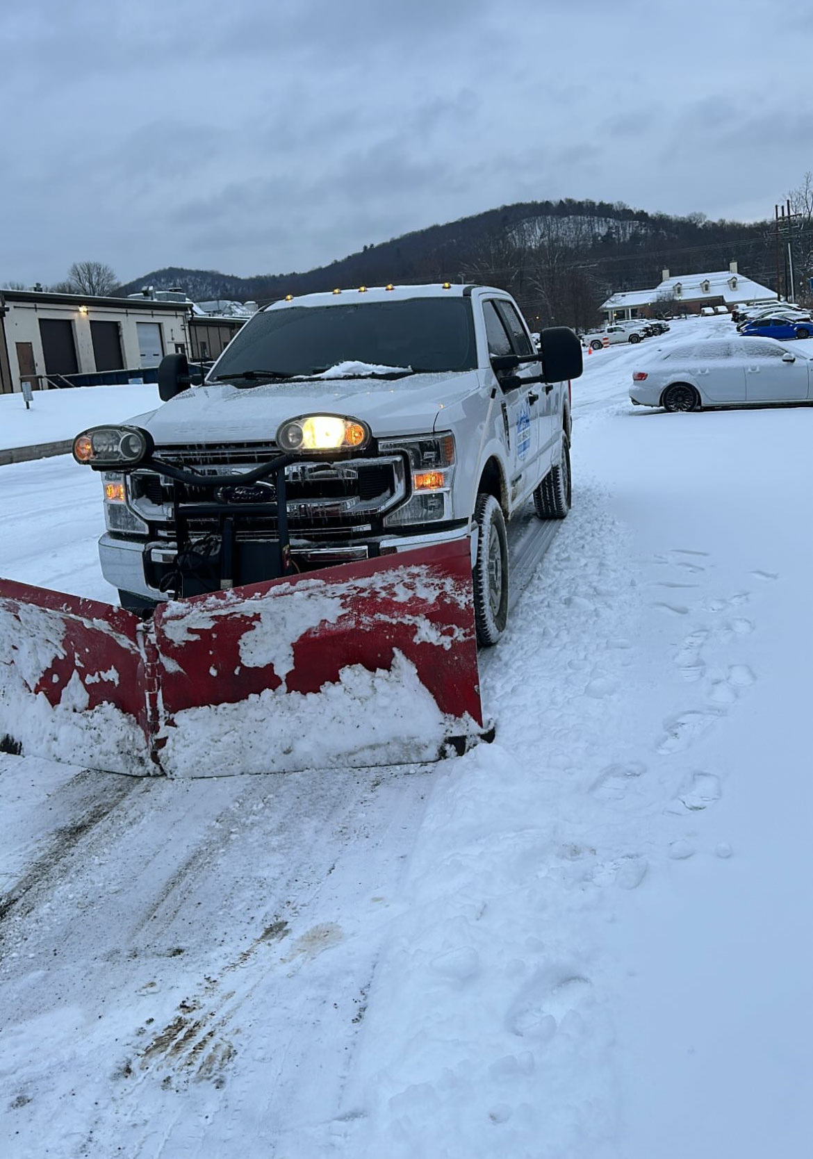 Plow truck with a red V-plow clearing deep snow from a retail parking lot