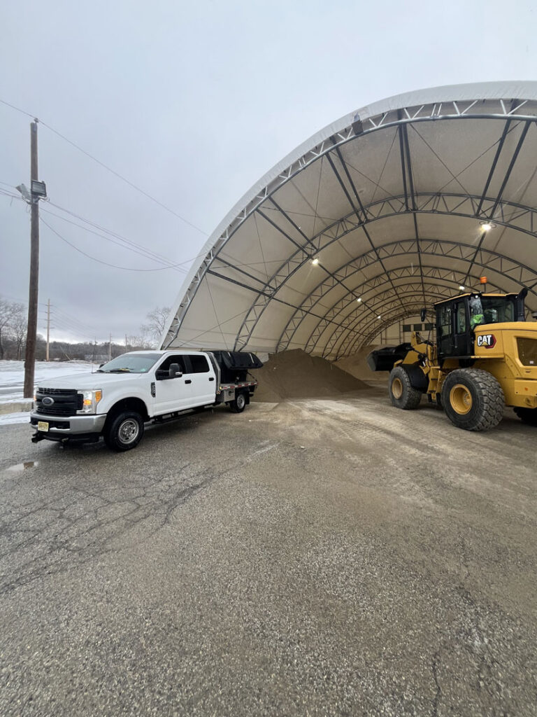 Wheel loader filling a JRW Services truck with salt at a hoop-style storage barn